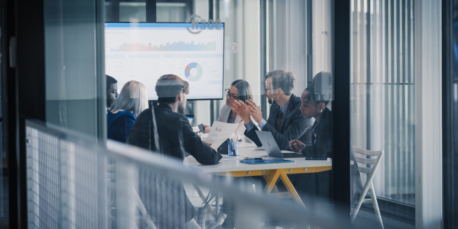 Líderes empresariais estão reunidos ao redor de uma mesa de conferência, analisando gráficos em uma tela grande, debatendo resultados, definindo prioridades e planejando os próximos passos, enquanto utilizam notebooks, relatórios impressos e anotações.