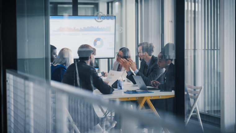 Líderes empresariais estão reunidos ao redor de uma mesa de conferência, analisando gráficos em uma tela grande, debatendo resultados, definindo prioridades e planejando os próximos passos, enquanto utilizam notebooks, relatórios impressos e anotações.